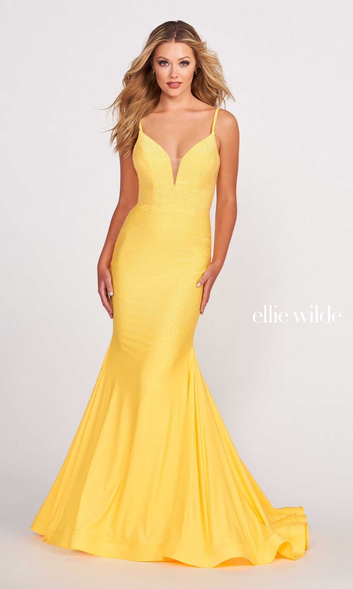 A woman wears the Ellie Wilde Beaded Mermaid Prom Dress with Cut Out, featuring a fitted yellow silhouette and deep V-neckline, standing against a plain light background. This gown radiates elegance and sophistication.